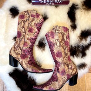 Könnte beinhalten: Ein Paar hohe Stiefel mit spitzer Zehenpartie und Schlangenmuster in Beige-, Burgunder- und Rosatönen. Die Stiefel haben schwarze Blockabsätze und sind auf einem weiß-schwarz gemusterten Teppich platziert. Der Text "THE MINI MAXI ARCHIVES" ist sichtbar.
