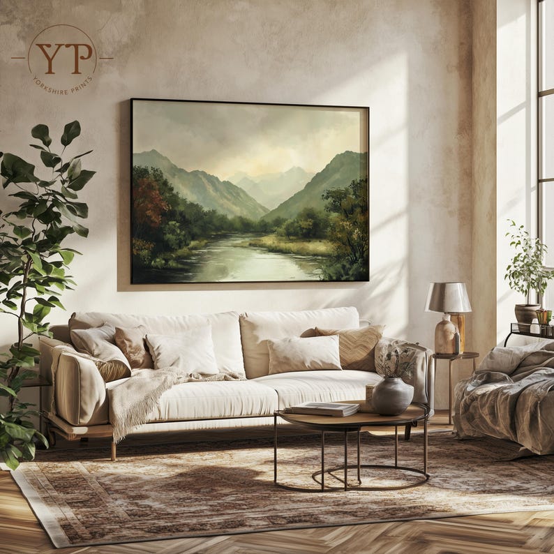 May include: A beige sofa with white throw pillows sits in a living room with a large framed landscape painting of a river and mountains. A small round coffee table with a book and a vase sits in front of the sofa. A rug with a brown and beige pattern covers the floor.