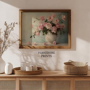 May include: A framed print of a vintage still life featuring a bouquet of pink roses in a white pitcher on a light blue background. The print is displayed on a wooden shelf with a wicker basket and white ceramic vases.