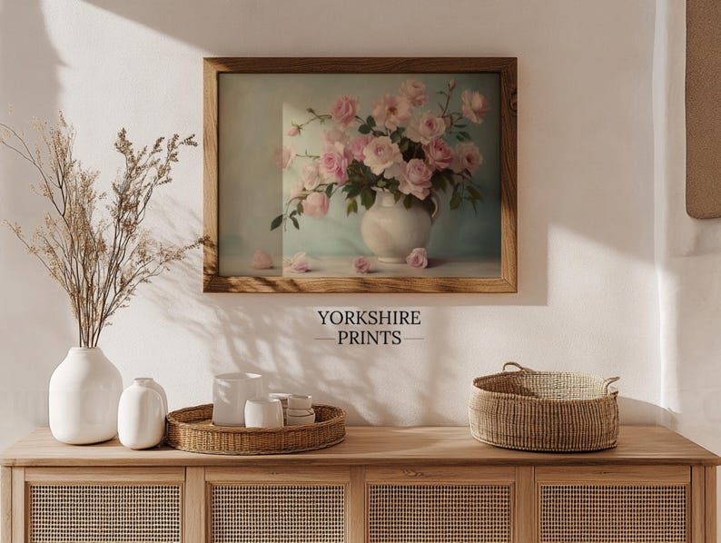 May include: A framed print of a vintage still life featuring a bouquet of pink roses in a white pitcher on a light blue background. The print is displayed on a wooden shelf with a wicker basket and white ceramic vases.