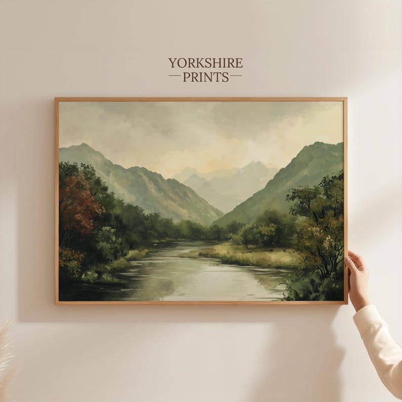 May include: A framed painting of a landscape with a river winding through a valley surrounded by green mountains. The sky is a soft gray with hints of pink and orange.