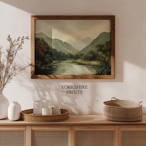 May include: A framed landscape painting of a river flowing through a valley with mountains in the background. The painting is hanging on a white wall above a wooden dresser with a wicker basket and vases.