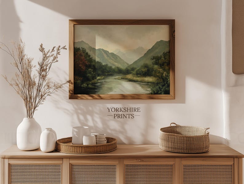 May include: A framed landscape painting of a river flowing through a valley with mountains in the background. The painting is hanging on a white wall above a wooden dresser with a wicker basket and vases.