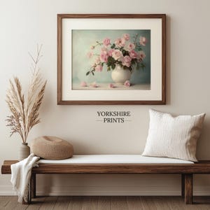May include: A framed print of a vintage still life featuring a white pitcher filled with pink roses against a light blue background. The print is displayed on a wooden bench with a throw blanket and a decorative pillow.