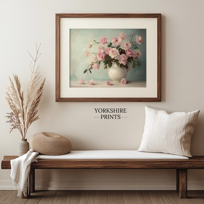 May include: A framed print of a vintage still life featuring a white pitcher filled with pink roses against a light blue background. The print is displayed on a wooden bench with a throw blanket and a decorative pillow.