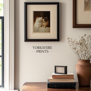 May include: A framed painting of two white kittens cuddling, hanging on a white wall with a wooden table and a vase of dried flowers in the foreground. The text "YORKSHIRE PRINTS" is printed below the painting.