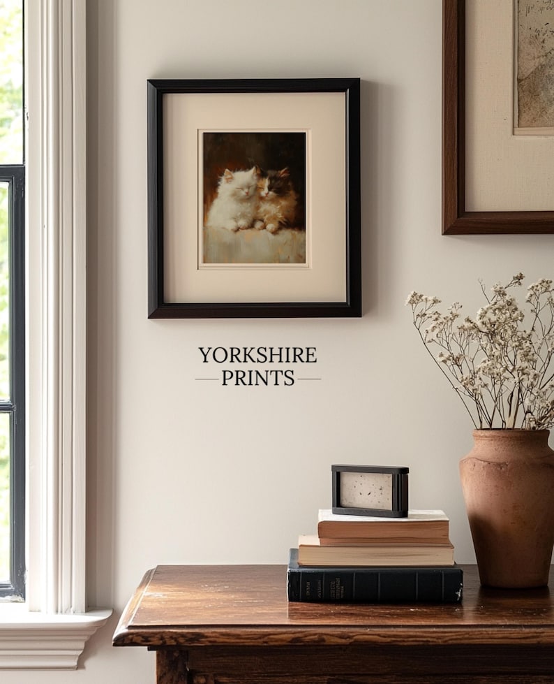 May include: A framed painting of two white kittens cuddling, hanging on a white wall with a wooden table and a vase of dried flowers in the foreground. The text "YORKSHIRE PRINTS" is printed below the painting.