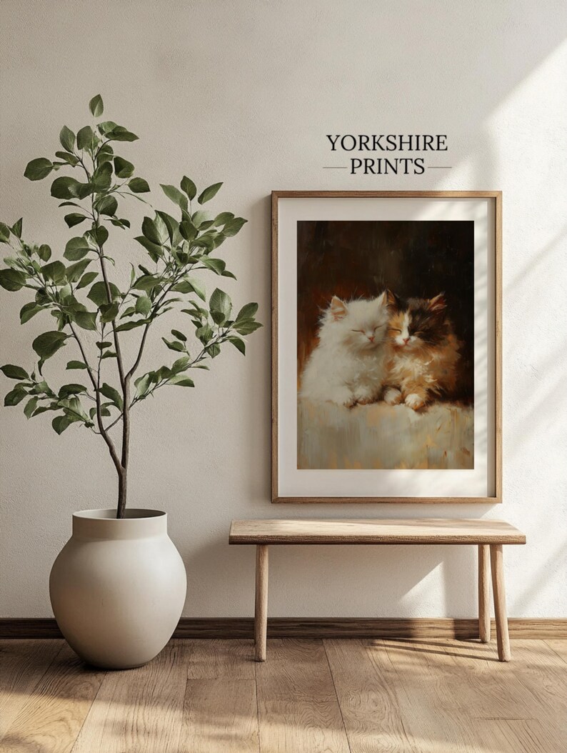 May include: A framed print of a painting depicting two cats cuddling together. The cats are white and brown with black markings. The print is set against a white wall with a potted plant and a wooden bench in the foreground. The text "YORKSHIRE PRINTS" is displayed above the print.