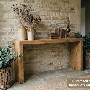 Peut inclure: Une console en bois rustique avec une finition naturelle, ornée d'objets décoratifs. La table est placée contre un mur en pierre et présente une lampe, des livres et des vases. Un panier en osier avec de la verdure est à gauche.