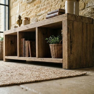 May include: A weathered wooden storage unit with three open sections. The unit is made of reclaimed wood and is placed against a stone wall. Books and a potted plant in a wicker basket are visible inside. A woven rug is in front of the unit.