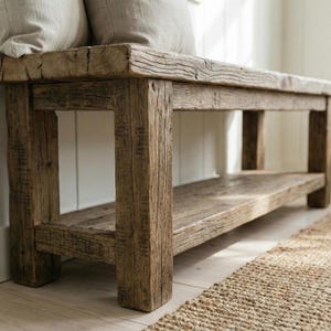 May include: A weathered wooden bench with a lower shelf. The bench is made of light brown wood with visible grain and knots. The design is simple and sturdy, suitable for a hallway or entryway. Two beige cushions rest on the bench.
