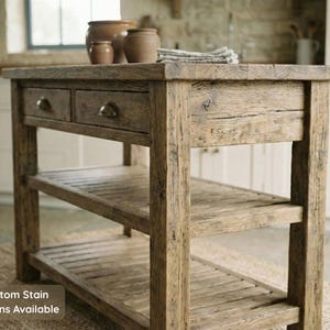 May include: A weathered wooden kitchen island with two drawers and two lower shelves. The island is made of reclaimed wood and has metal handles. The text "Custom Stain Options Available" is visible.