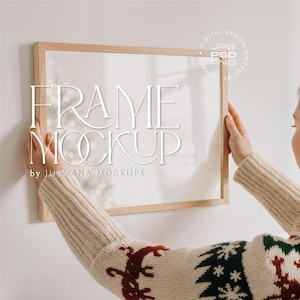 Puede incluir: Una maqueta de marco rectangular de madera con el texto "FRAME MOCKUP" en blanco. El marco lo sostiene una persona que lleva un suéter beige con un estampado festivo. La imagen incluye un logotipo de diseño digital.