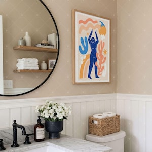 Puede incluir: Una l&aacute;mina de arte enmarcada que muestra una figura humana azul rodeada de formas abstractas en naranja, amarillo y azul claro sobre un fondo crema. La obra est&aacute; expuesta en un marco de madera clara sobre una pared beige en un ba&ntilde;o.