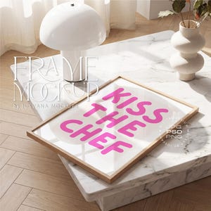 Puede incluir: Una impresión enmarcada con el texto "KISS THE CHEF" en letras de color rosa brillante. La impresión está en un marco de madera clara y descansa sobre una mesa de mármol. Una lámpara y un jarrón blancos también están sobre la mesa, con un suelo de madera en el fondo.
