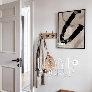 Puede incluir: Una impresión artística abstracta enmarcada con pinceladas negras sobre un fondo beige. Un perchero de madera con ganchos negros sostiene un abrigo de color claro y una bolsa de paja. Se muestra el texto "FRAME MOCKUP".