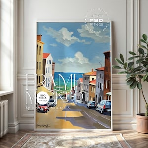 Puede incluir: Una impresión artística enmarcada que presenta una colorida escena callejera con edificios, una carretera y coches. La obra de arte representa una ciudad costera con un cielo azul y el mar. El marco es blanco y la impresión se muestra en una pared con una planta.