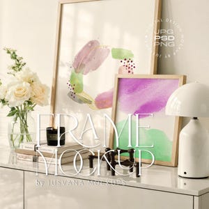 Puede incluir: Maqueta con dos impresiones artísticas enmarcadas con diseños abstractos en tonos morados, verdes y dorados. Una lámpara blanca, una vela y libros están sobre un mueble blanco. El texto "FRAME MOCKUP" está superpuesto.