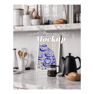 Puede incluir: Un marco rectangular blanco que muestra una ilustración gráfica azul y beige de tazas de café y pasteles sobre una superficie a cuadros. El marco está colocado sobre una encimera de mármol en una cocina con un hervidor negro y una jarra de cristal.