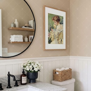 Puede incluir: Un ba&ntilde;o con un espejo redondo de marco negro que refleja estantes de madera con toallas blancas, un retrato enmarcado de una persona con una flor amarilla, un lavabo de m&aacute;rmol con grifo negro y una cesta de mimbre sobre el inodoro.