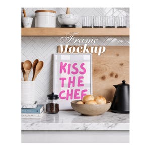 Puede incluir: Un marco de fotos blanco con el texto rosa KISS THE CHEF, exhibido en una encimera de cocina con un cuenco de panecillos, un hervidor negro, un tarro de cristal con granos de café, cucharas de madera y libros apilados ante una pared de azulejos.