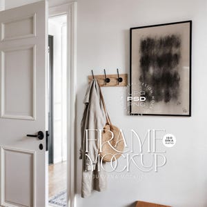 Puede incluir: Escena interior de tonos neutros con una puerta blanca, un perchero de madera con ganchos negros y una obra de arte abstracta enmarcada. Un abrigo beige y un bolso de paja cuelgan del perchero. El texto "FRAME MOCKUP" es visible.