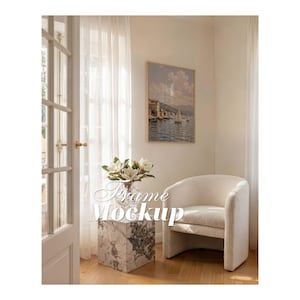 Op de afbeelding: Een rechthoekige houten fotolijst met een print van een kustlandschap met zeilboten op het water, bevestigd aan een lichte muur naast een witte, getextureerde fauteuil, een marmeren kubustafel en een glazen vaas met witte bloemen.