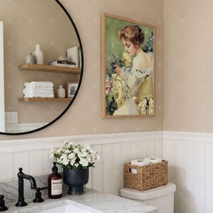 Puede incluir: Una impresi&oacute;n art&iacute;stica rectangular enmarcada que muestra el retrato de una persona con cabello casta&ntilde;o sosteniendo una flor amarilla, exhibida en un ba&ntilde;o con un espejo redondo de marco negro, un tocador de m&aacute;rmol y una cesta tejida.