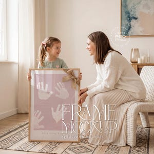 Puede incluir: Un marco con huellas de manos sobre un fondo rosa, atado con un lazo de arpillera. El marco lo sostienen una niña y una mujer. Las palabras "FRAME MOCKUP" se muestran en el marco.