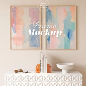 May include: Two framed abstract watercolor paintings with pastel colors hang above a white geometric cabinet. The cabinet holds books, orange bookends, a white bowl, sunglasses, and keys. The text "Frame Mockup" is overlaid on the paintings.