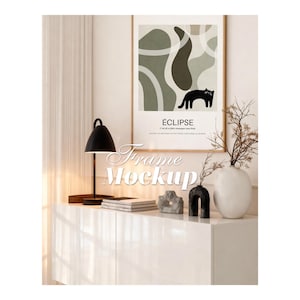Puede incluir: Una lámina enmarcada titulada ÉCLIPSE con formas orgánicas abstractas en verde salvia y blanco junto a la silueta de un gato negro. La obra se exhibe sobre un mueble blanco con una lámpara de escritorio negra, libros y dos jarrones.