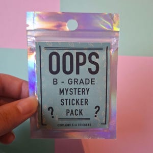 Peut inclure: Un emballage holographique irisé contenant un "OOPS B-GRADE MYSTERY STICKER PACK". Le paquet d'autocollants a un fond turquoise avec du texte noir et des points d'interrogation. Le paquet contient 5 à 6 autocollants.