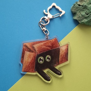 Puede incluir: Un llavero acrílico transparente con un gato de dibujos animados asomando por una caja de cartón marrón. El gato tiene ojos verdes y pelaje negro. El llavero tiene un cierre plateado.