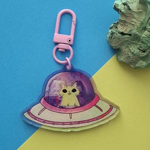 Puede incluir: Un llavero acrílico transparente con un gato de dibujos animados en un OVNI. El OVNI es blanco con detalles en rosa y un contorno rosa. El llavero tiene un cierre y un anillo rosas. El fondo es azul y amarillo.