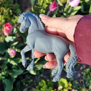 May include: A gray horse figurine with a detailed mane and tail, held in a hand. The horse is posed with one leg raised, showcasing its sculpted muscles. The background features green foliage and colorful flowers.