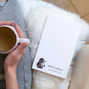 May include: A white notepad with a cartoon penguin wearing glasses and the text "DON'T FORGET TO REMEMBER THIS." A person holds a white mug with a brown interior. The person is wearing ripped blue jeans.