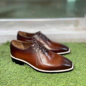 Puede incluir: Un par de zapatos marrones de cuero con cordones y un borde blanco alrededor de la suela. Los zapatos est&aacute;n sobre una superficie de hierba verde.