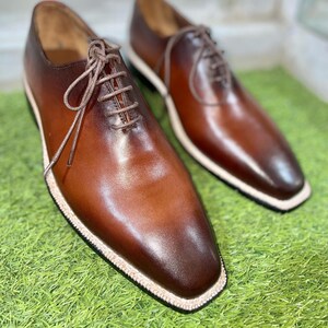 Puede incluir: Un par de zapatos marrones de cuero con cordones y un borde blanco alrededor de la suela. Los zapatos est&aacute;n sobre una superficie de hierba verde.