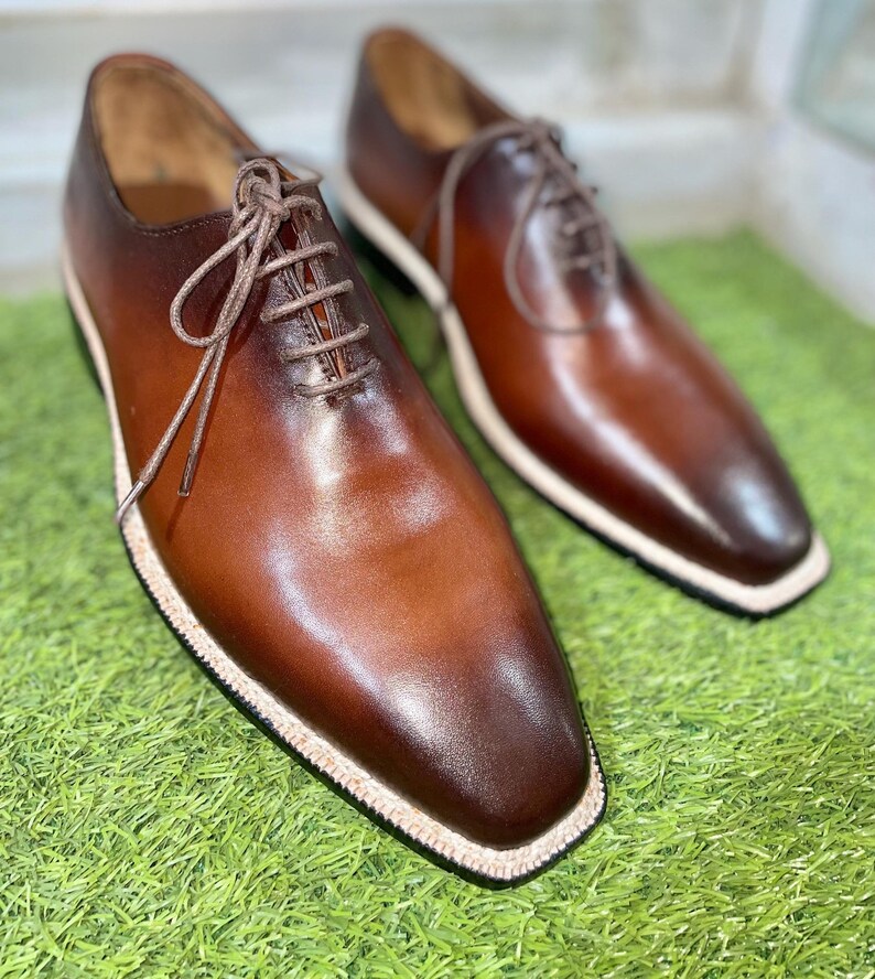 Puede incluir: Un par de zapatos marrones de cuero con cordones y un borde blanco alrededor de la suela. Los zapatos est&aacute;n sobre una superficie de hierba verde.