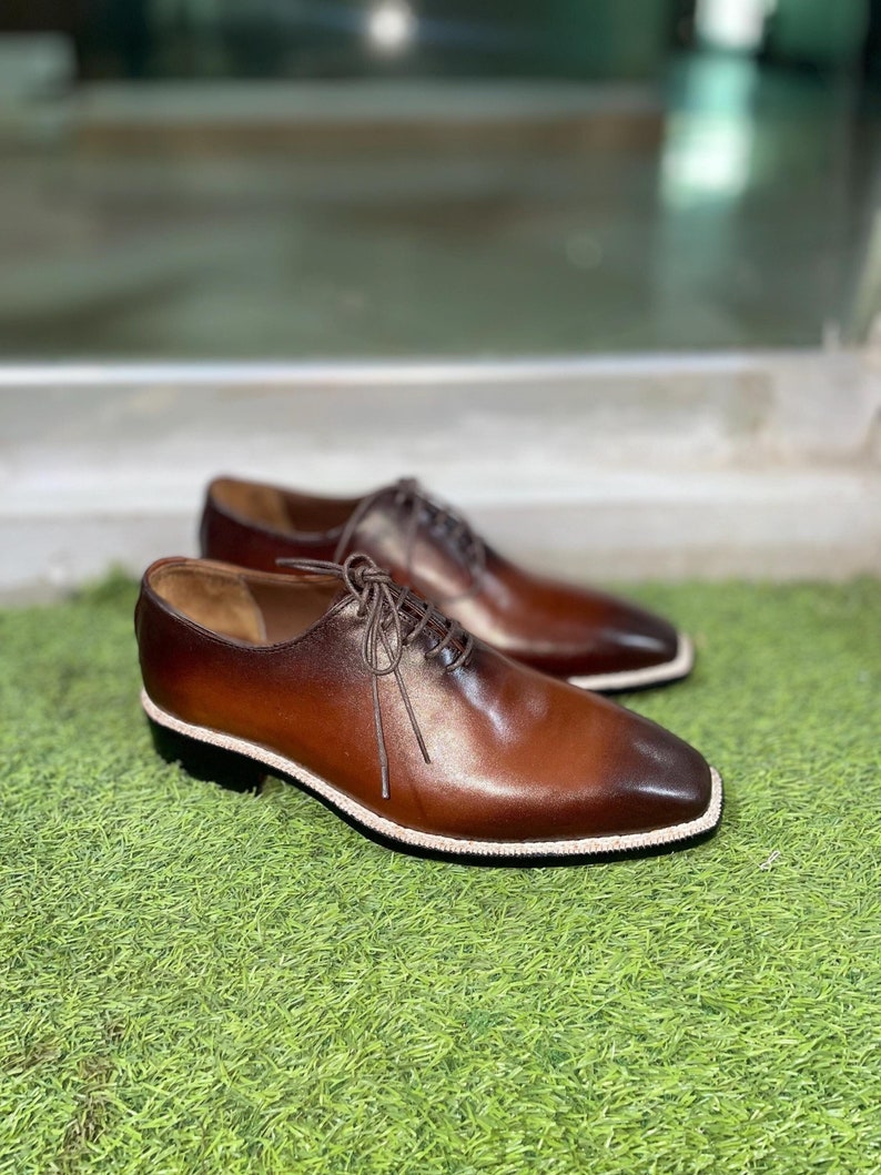 Puede incluir: Un par de zapatos marrones de cuero con cordones y un borde blanco alrededor de la suela. Los zapatos est&aacute;n sobre una superficie de hierba verde.