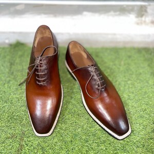 Puede incluir: Un par de zapatos marrones de cuero con cordones y costuras blancas alrededor de la suela. Los zapatos est&aacute;n sobre una superficie de c&eacute;sped artificial verde.