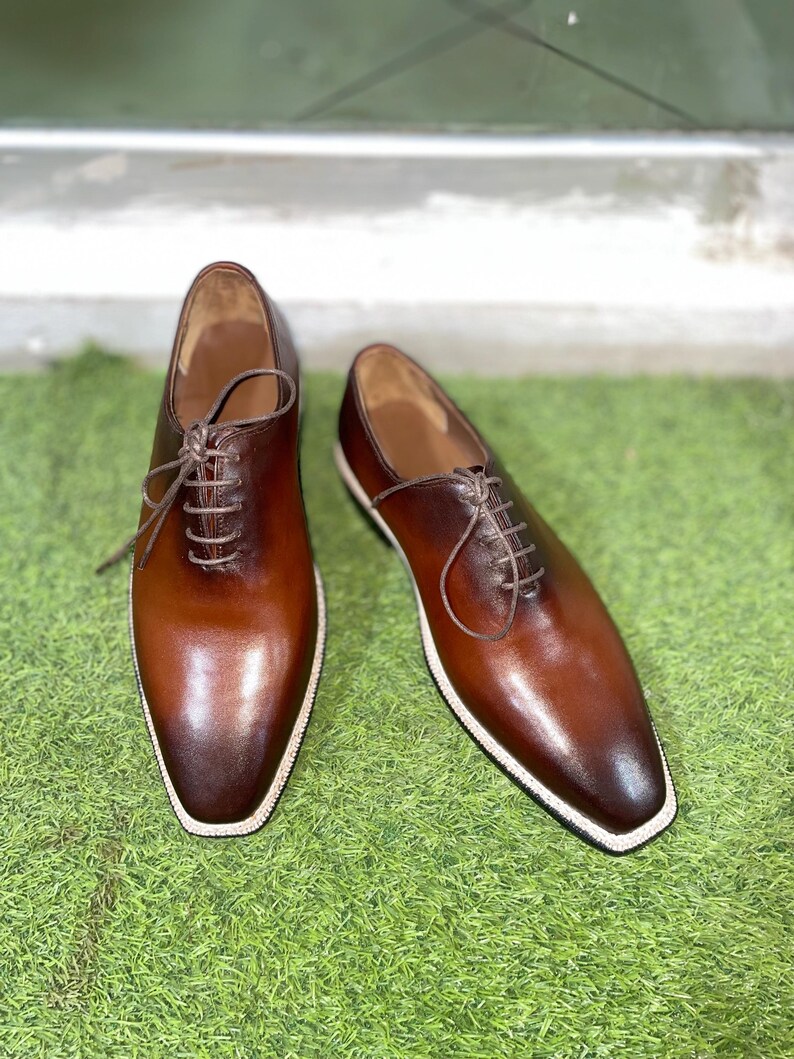 Puede incluir: Un par de zapatos marrones de cuero con cordones y costuras blancas alrededor de la suela. Los zapatos est&aacute;n sobre una superficie de c&eacute;sped artificial verde.