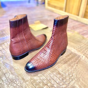 Puede incluir: Un par de botas de cuero marrón con estampado de piel de serpiente. Las botas tienen una punta puntiaguda y una cremallera en el lateral. Las botas están sobre una superficie de cuero marrón.