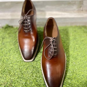 Puede incluir: Un par de zapatos de cordones de cuero marr&oacute;n con punta puntiaguda y un borde blanco alrededor de la suela.