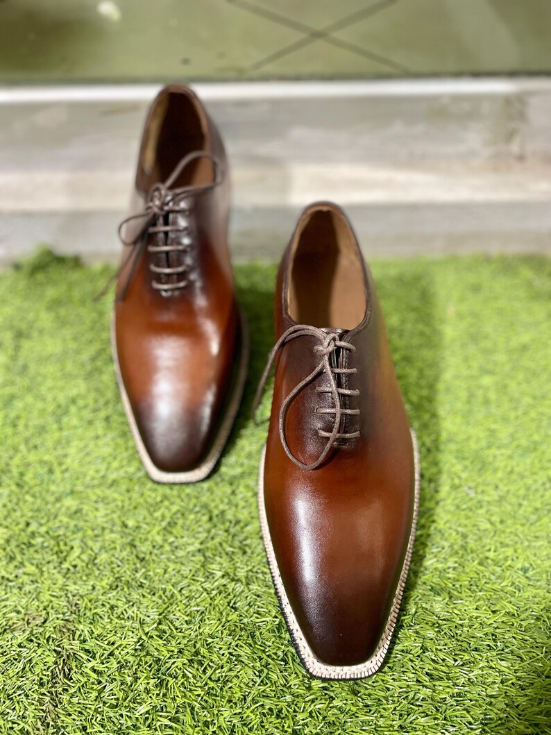 Puede incluir: Un par de zapatos de cordones de cuero marr&oacute;n con punta puntiaguda y un borde blanco alrededor de la suela.