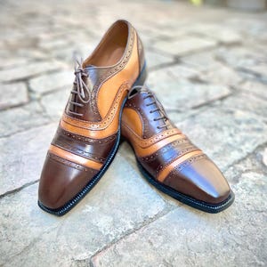 May include: A pair of brown and tan leather oxford shoes with brogue detailing. The shoes have a lace-up closure and a rounded toe.