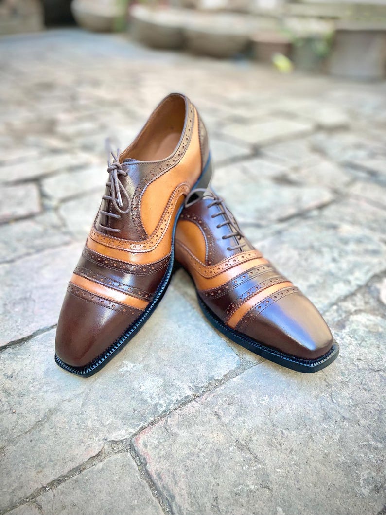 May include: A pair of brown and tan leather oxford shoes with brogue detailing. The shoes have a lace-up closure and a rounded toe.