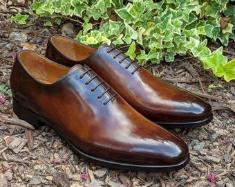 Puede incluir: Un par de zapatos de vestir de cuero marrón con cordones y acabado brillante. Los zapatos tienen punta puntiaguda y tacón bajo.