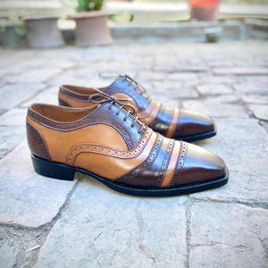 May include: A pair of brown and tan leather lace-up shoes with a brogue design. The shoes have a black sole and are sitting on a stone surface.