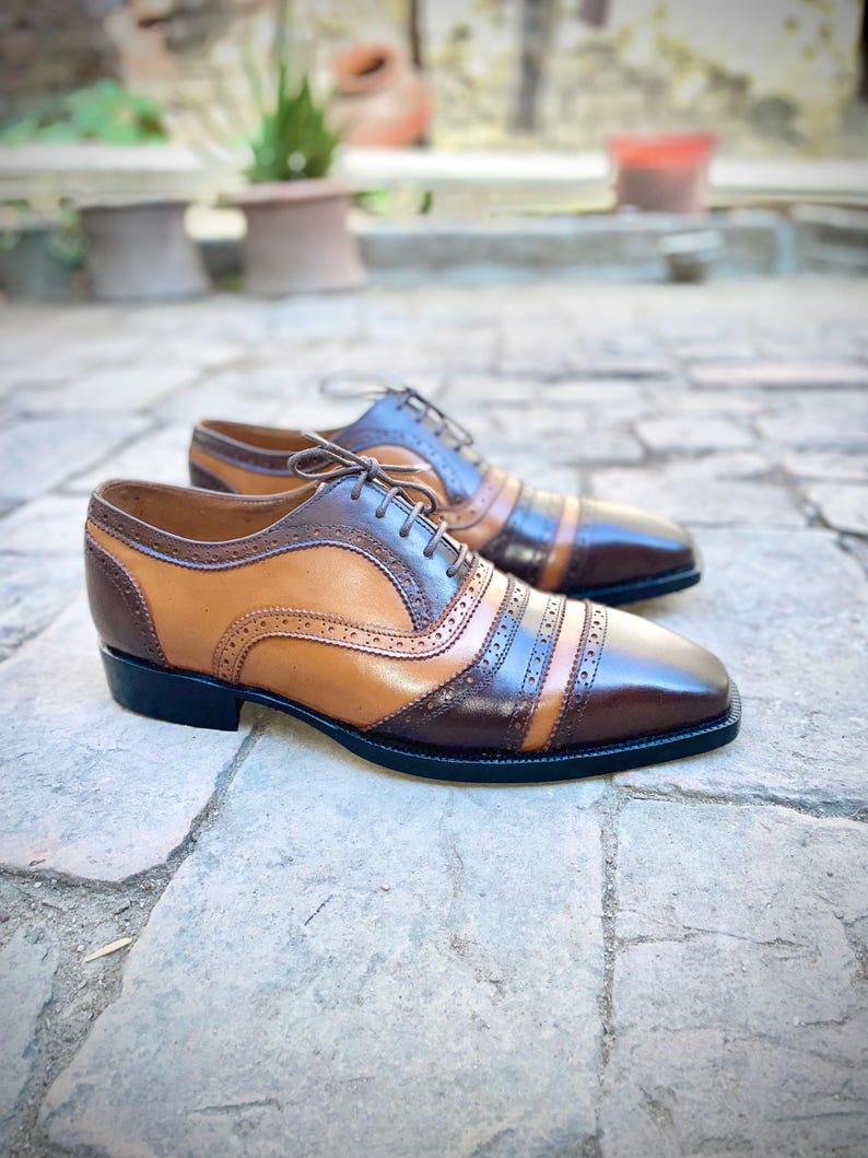 May include: A pair of brown and tan leather lace-up shoes with a brogue design. The shoes have a black sole and are sitting on a stone surface.